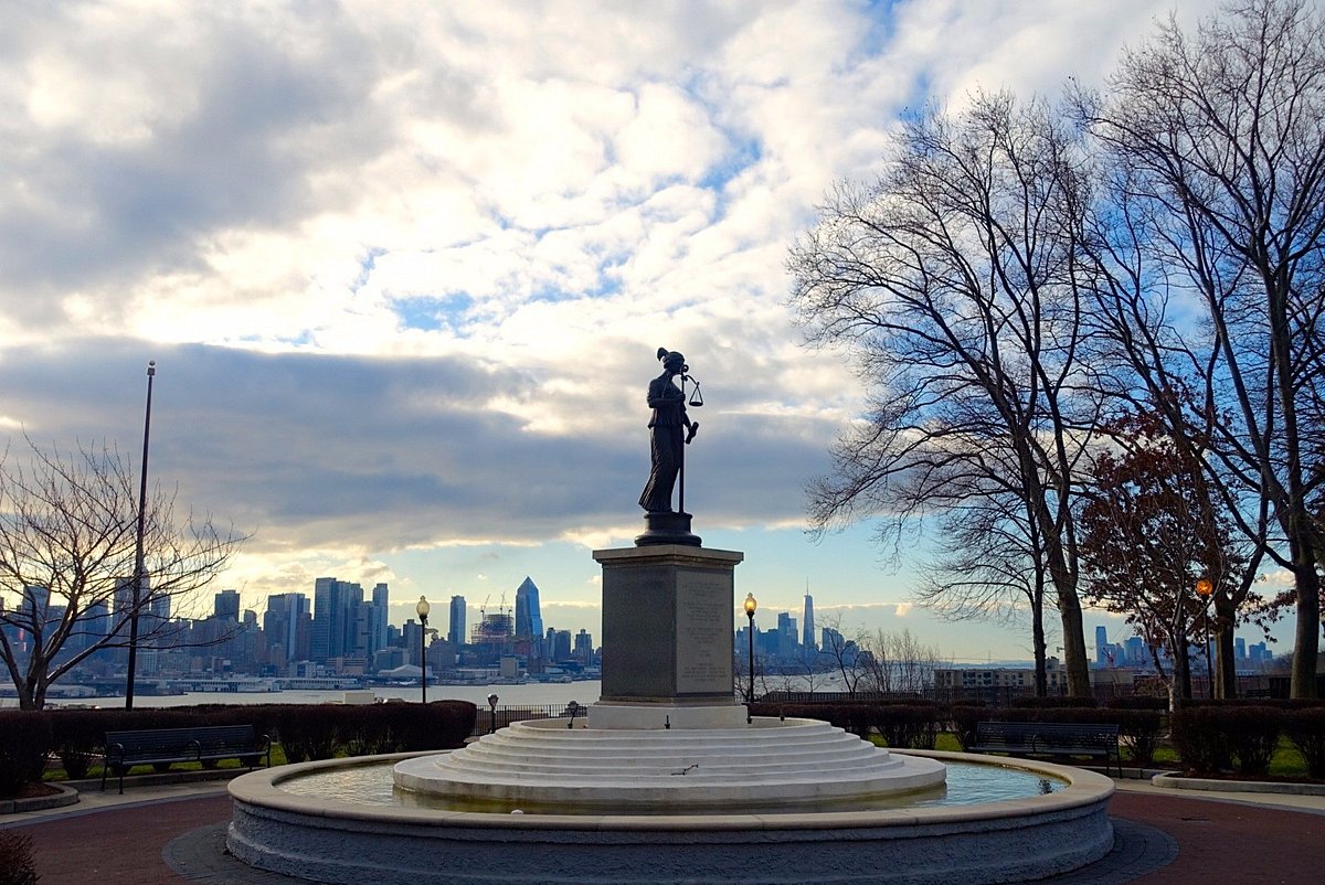 Statue of Justice with scales in a park setting, overlooking the skyline of Jersey City and the Hudson River, surrounded by trees and a fountain, representing a local landmark near Uforia Dispensary in West New York, NJ.