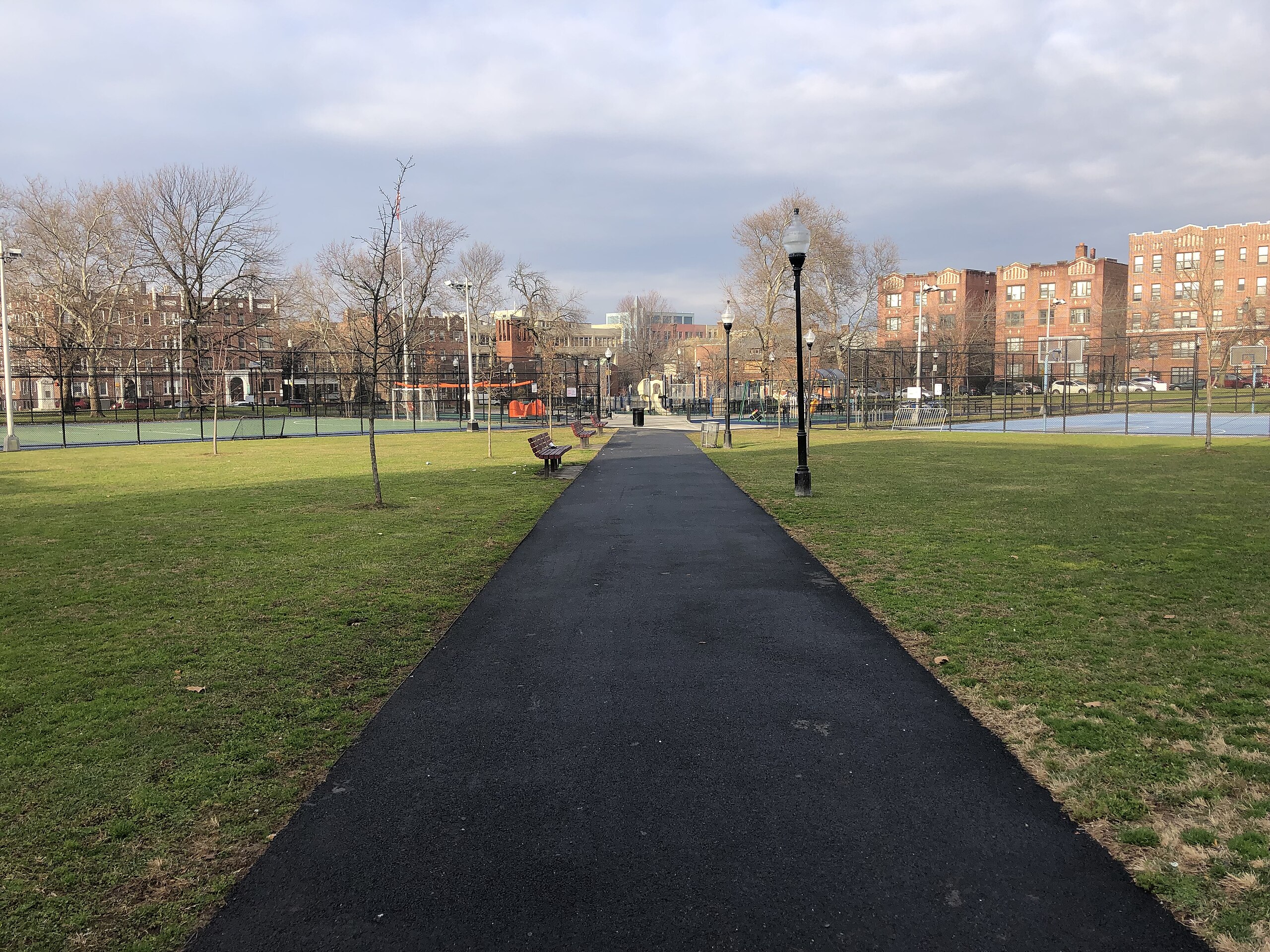 Pathway through Audubon Park in Jersey City, lined with benches and lampposts, leading to sports courts and residential buildings.