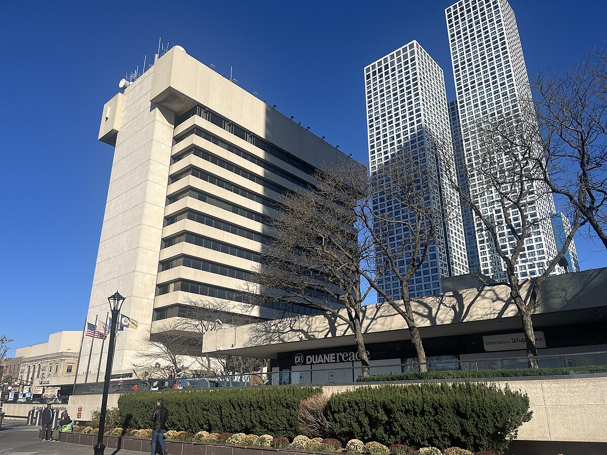 Journal Square area in Jersey City, featuring Uforia Dispensary's modern building alongside high-rise towers and nearby retail establishments, with clear blue sky and pedestrians.