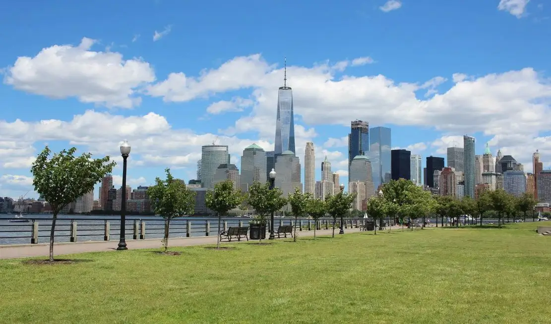View of Liberty State Park with green grass and trees, overlooking the Jersey City skyline featuring One World Trade Center and other skyscrapers against a blue sky with clouds, highlighting proximity to Uforia Dispensary.