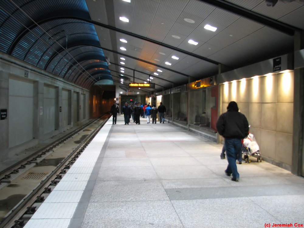 Interior view of a modern transit station with passengers walking along the platform, train tracks visible, and overhead signage indicating Bergenline Avenue Station.