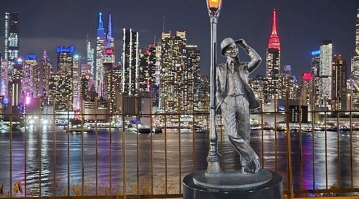 Statue of a man in a hat leaning against a lamppost with the New York City skyline illuminated at night in the background, representing the vibrant community near Uforia Dispensary in Weehawken, NJ.