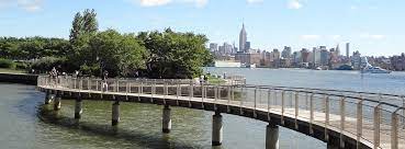 Scenic view of a waterfront walkway leading to the New York City skyline, featuring the Empire State Building, with lush greenery and a clear blue sky, highlighting nearby landmarks for Uforia Dispensary in Hoboken, NJ.