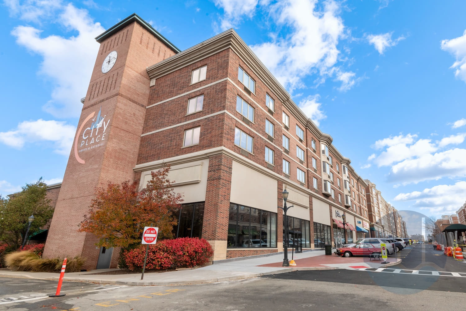 City Place at the Promenade, a multi-story brick building featuring shops and restaurants, located in Edgewater, NJ, with clear blue skies and autumn foliage.
