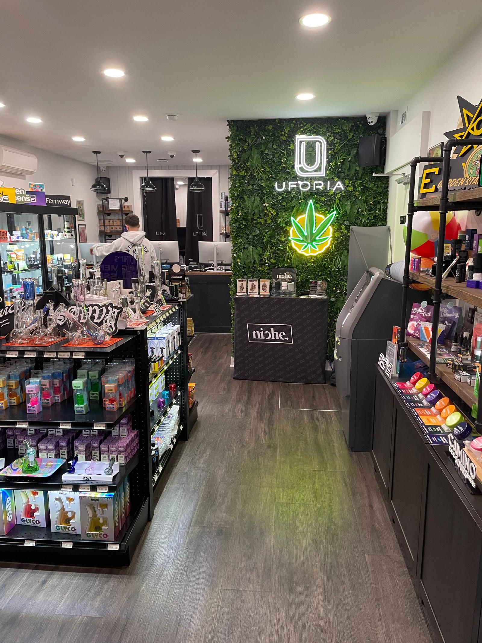 Uforia Dispensary interior showcasing cannabis products, vibrant green wall with Uforia logo, display shelves filled with smoking accessories and vape products, emphasizing a welcoming atmosphere for customers in Jersey City, NJ.