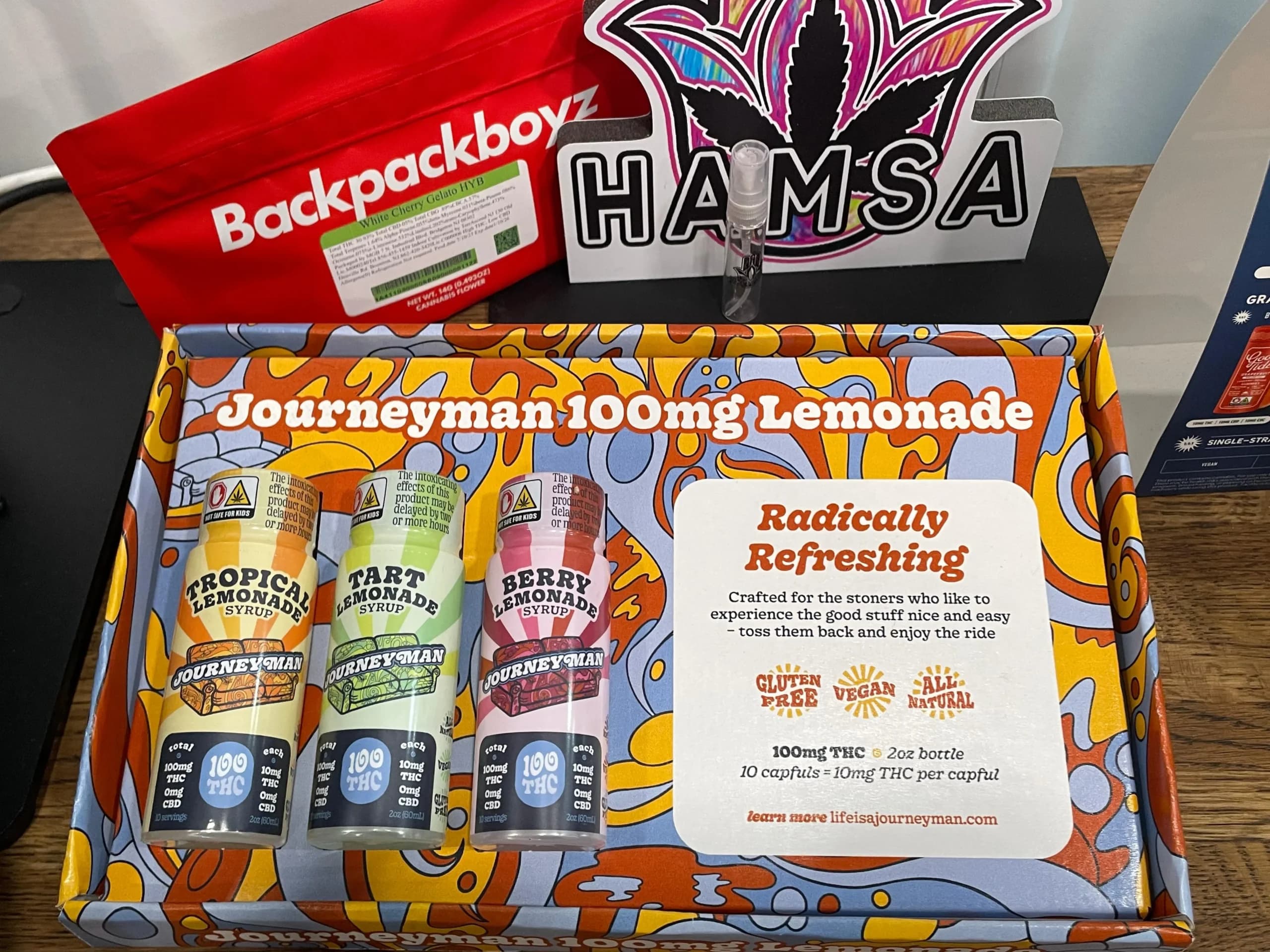 Craft cannabis beverage box featuring Journeyman 100mg lemonade syrups in Tropical, Tart, and Berry flavors, with a colorful design and product information highlighting gluten-free, vegan, and all-natural attributes, set against a backdrop of cannabis-themed decor.
