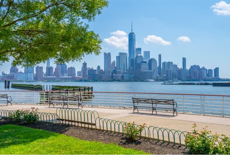 View of the Hudson River Waterfront Walkway with benches and greenery, showcasing the Manhattan skyline, relevant for visitors seeking directions to Uforia Dispensary in Jersey City.