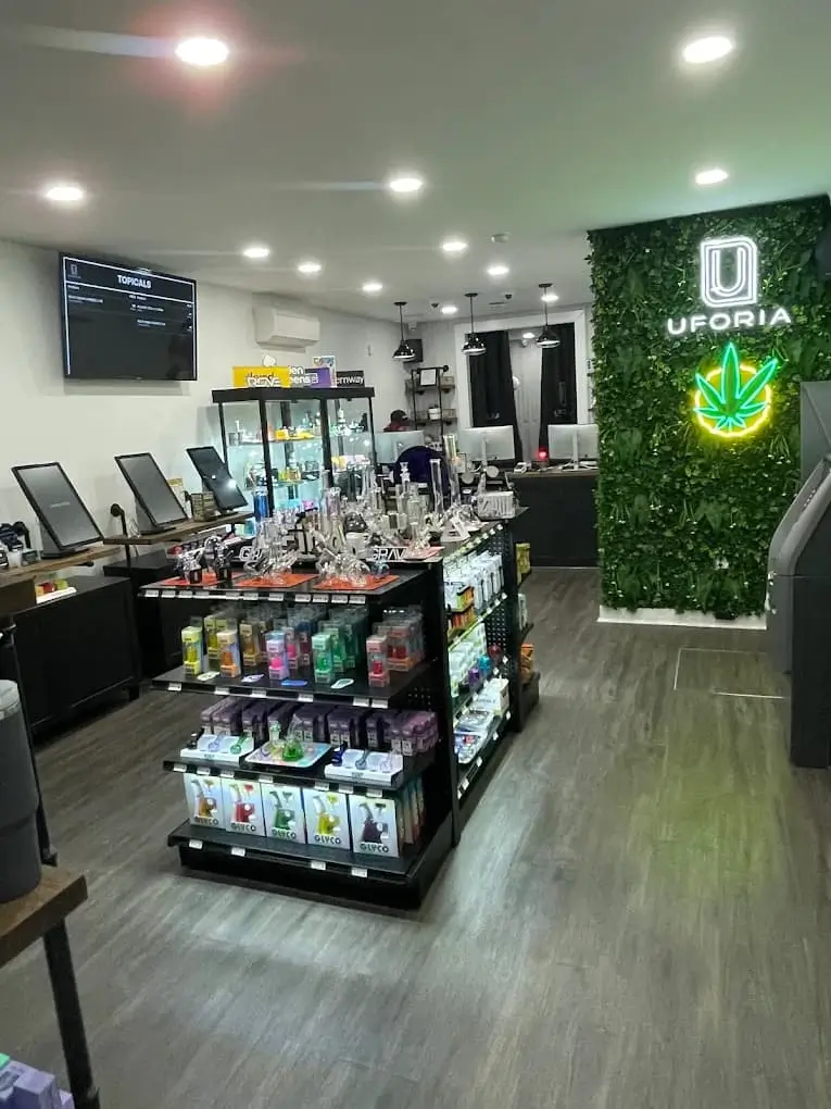 Uforia Dispensary interior showcasing cannabis products, display shelves with edibles, vapes, and accessories, vibrant green wall with Uforia logo and neon cannabis leaf, modern retail environment in Jersey City.