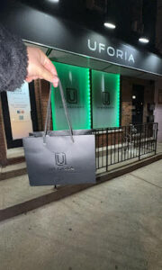 Hand holding a black shopping bag outside UFORIA storefront with green lighting